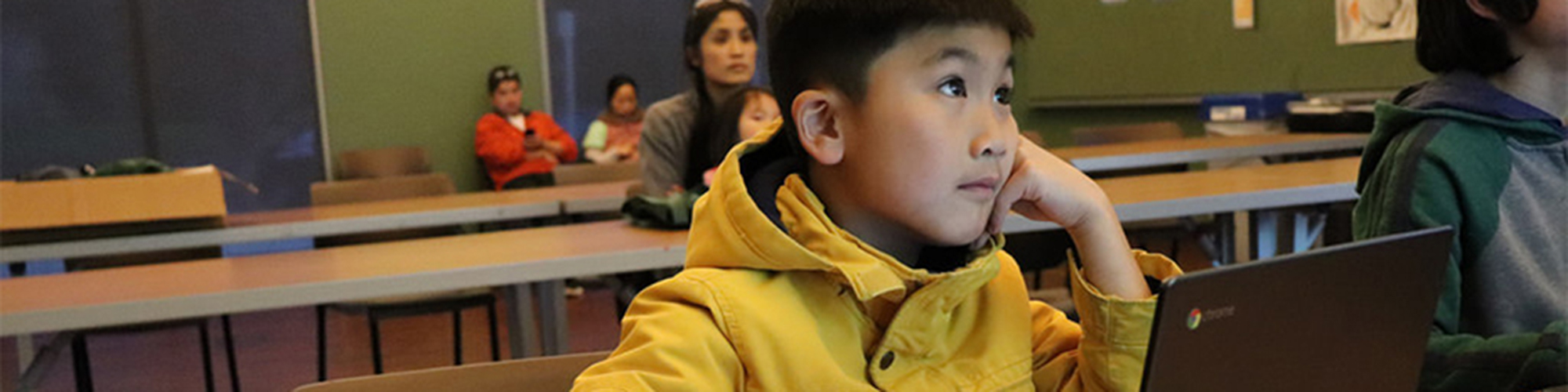 A young boy in a yellow jacket sits behind a computer.